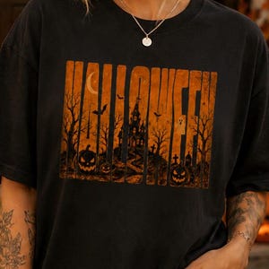 May include: A black short-sleeve t-shirt featuring a rectangular orange graphic print with the word HALLOWEEN in bold letters, a haunted house, bare trees, a crescent moon, and jack-o'-lanterns. The shirt is paired with light blue distressed jeans.