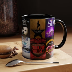 Pode incluir: Uma caneca de cerâmica preta com uma colagem de cartazes de musicais da Broadway, incluindo The Phantom of the Opera, Hamilton, Six, Dear Evan Hansen, Moulin Rouge e Six. A caneca tem uma asa e interior pretos com designs gráficos coloridos.