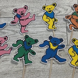 May include: A set of twelve dancing bear cupcake toppers in various colors including blue, pink, yellow, green, and orange. Each bear figure is attached to a thin wooden pick, featuring a distinct color combination with contrasting neck ruffs.