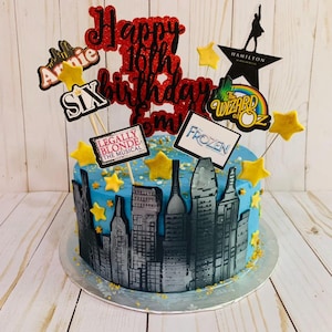 May include: A round blue cake decorated with a black and grey city skyline silhouette around the base. The top features glittery red text reading Happy 16th Birthday, yellow star accents, and various musical theater themed toppers including Annie, Six, Hamilton, The Wizard of Oz, Legally Blonde, and Frozen.