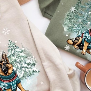Peut inclure: Deux sweats à capuche avec une scène d'hiver représentant un berger allemand portant un pull de fête. Le chien est assis dans une forêt enneigée avec des sapins. Les sweats à capuche sont disponibles en beige et vert olive.