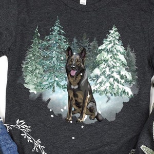 Op de afbeelding: Een donkergrijs longsleeve t-shirt met een wintersc&egrave;ne met een Duitse herder die in een besneeuwd bos staat. De hond is bruin en zwart met een witte borst en poten. De bomen zijn groen en bedekt met sneeuw. De achtergrond is een lichtblauwe aquarelwas.
