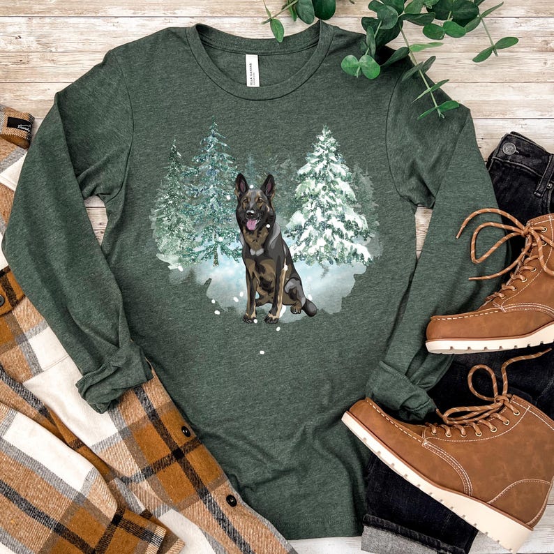 Op de afbeelding: Een groen shirt met lange mouwen met een afbeelding van een Duitse herder die in een besneeuwd bos zit. De hond is bruin en zwart met een witte borst.