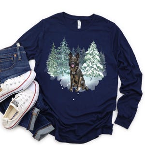 Op de afbeelding: Een marineblauw longsleeve t-shirt met een afbeelding van een Duitse herder in een besneeuwd bos. De hond is bruin en zwart en zit in de sneeuw.