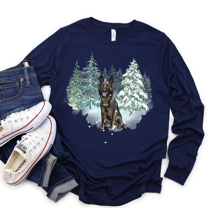 Op de afbeelding: Een marineblauw longsleeve t-shirt met een afbeelding van een Duitse herder in een besneeuwd bos. De hond is bruin en zwart en zit in de sneeuw.