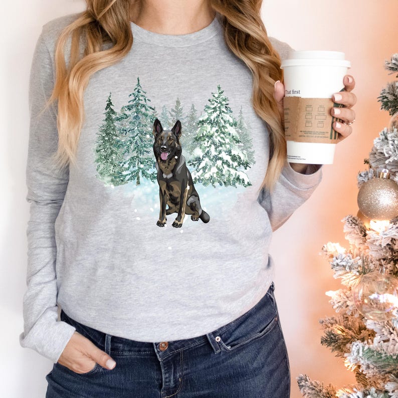 Op de afbeelding: Een grijs longsleeve shirt met een afbeelding van een Duitse herder die in een besneeuwd bos zit. De hond is bruin en zwart met een witte borst en poten.