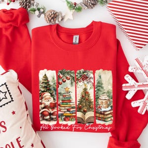 May include: Red Christmas sweatshirt with a graphic of a stack of books, a Christmas tree, a cup of coffee, and Santa Claus reading a book. The text "All Booked For Christmas" is printed below the graphic.