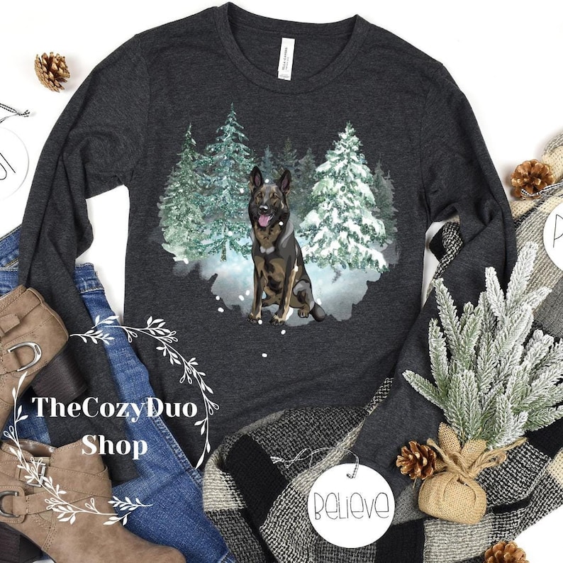 Op de afbeelding: Een donkergrijs longsleeve t-shirt met een wintersc&egrave;ne met een Duitse herder die in een besneeuwd bos staat. De hond is bruin en zwart met een witte borst en poten. De bomen zijn groen en bedekt met sneeuw. De achtergrond is een lichtblauwe aquarelwas.