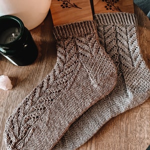 Puede incluir: Un par de calcetines de punto marrón con un diseño de hojas, exhibidos en hormas de madera. Los calcetines tienen un puño acanalado y un diseño calado detallado. En el fondo, una vela y un pequeño objeto decorativo.