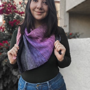 Puede incluir: Una mujer con una camisa negra de manga larga y pantalones vaqueros azules. Está sosteniendo una chal de punto degradado en morado, rosa y gris.