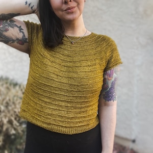 Puede incluir: Un top de punto amarillo mostaza con mangas cortas y cuello redondo. El top está hecho de un hilo suave y texturizado y tiene un corte ligeramente recortado.