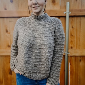 Puede incluir: Un suéter de punto marrón con cuello alto y mangas largas. El suéter está hecho de un hilo suave y texturizado y tiene un ajuste holgado.