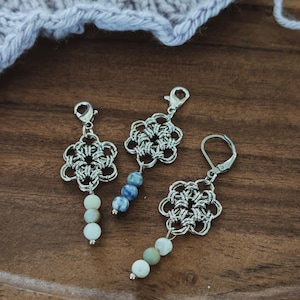 May include: Three silver-toned floral-shaped charms with beaded accents. Two charms have lobster clasps, and one has a lever-back earring hook. The beads are in shades of white, blue, and green. The background is a wooden surface with a knitted item.