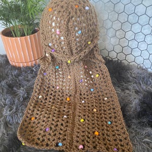 May include: A brown crocheted hood with colorful beaded accents. The hood is made of a textured yarn and features a decorative open weave design. The hood is a unique accessory.
