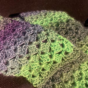 May include: A crocheted textile featuring a repeating granny square stitch pattern. The yarn displays a variegated colour gradient transitioning through shades of deep purple, slate blue, forest green, and bright lime green in a continuous sequence.