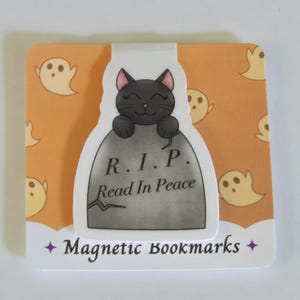 Puede incluir: Un gato negro con orejas rosas asoma por detrás de una lápida gris con el texto "R.I.P. Read In Peace". El fondo es naranja con un patrón de fantasmas blancos. El texto "Magnetic Bookmarks" está en la parte inferior de la imagen.