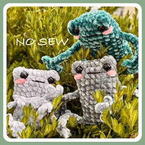 May include: Three plush frog toys made of soft, textured yarn in shades of teal, light grey, and dark grey. Each frog features black plastic eyes, pink stitched cheeks, and a simple horizontal mouth. The text NO SEW is visible in white.