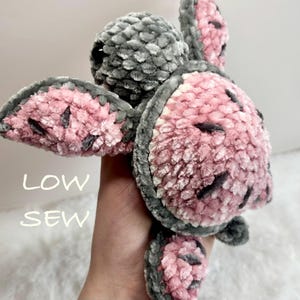 May include: A plush turtle toy crafted from soft, fuzzy yarn in shades of dusty pink and charcoal gray. The turtle features a rounded shell, four flippers, and a head with a dark eye. The text "LOW SEW" is visible on the left side of the image.