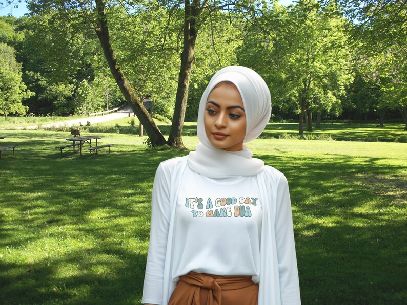 May include: A white long-sleeved shirt with the text "It's a good day to make dua" in colourful letters. The person is wearing a white hijab and brown trousers. The background is a green grassy field with trees.
