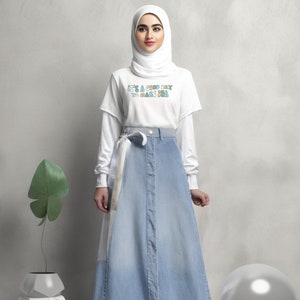 May include: A woman wearing a white t-shirt with the text "It's a good day to make you smile" in colourful letters, a white headscarf, and a light blue denim skirt with a white belt.