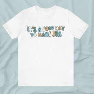 May include: White t-shirt with the text "It's a good day to make dua" in colourful, retro-style lettering.