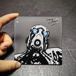 May include: Square glass coaster with a black and white illustration of a character with blue eyes and a mask. The artwork is set against a grey gradient background, with the artist's signature in the upper right corner.