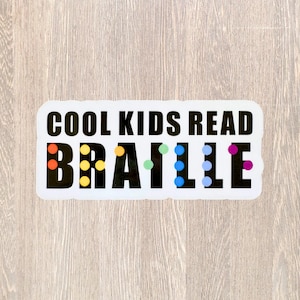 May include: A rectangular sticker with the text "COOL KIDS READ BRAILLE" in black. The word "BRAILLE" has colorful dots representing the braille letters. The sticker is on a light wood grain background.