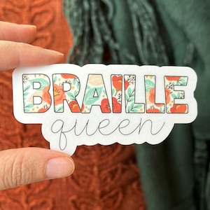 May include: A white sticker with the words "Braille queen" in black script. The word "Braille" is in large, bold letters and is filled with a floral pattern in shades of red, green, and white.