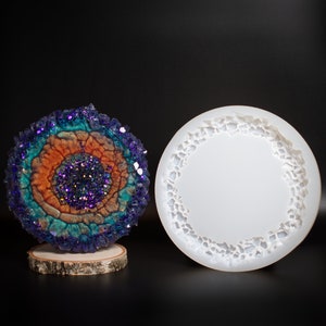 May include: A colorful geode art piece with a vibrant display of purple, teal, and orange hues, mounted on a wooden base. Next to it is a white, circular silicone mold with a textured edge, perfect for resin art projects.
