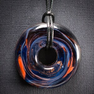 May include: A glass pendant necklace with a swirling pattern of blue, orange, and black. The pendant is shaped like a donut and hangs from a black cord.