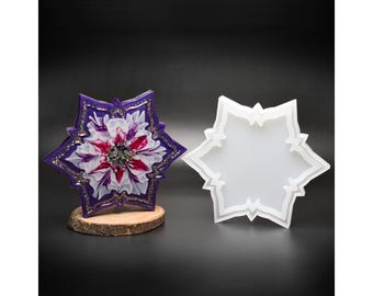 Può includere: Un sottobicchiere decorativo in resina con un design floreale nei toni del viola, bianco e rosso, impreziosito da glitter dorati. Accanto, uno stampo in silicone bianco con la stessa forma a stella. Il sottobicchiere è esposto su una fetta di legno.