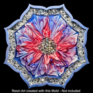 Op de afbeelding: Een harskunstwerk met een bloemmotief in rood, blauw en wit, met een sprankelend centrum. Het kunstwerk is geplaatst in een blauwe, sierlijke lijst, omgeven door een rand van heldere, reflecterende elementen. De algemene vorm is een gestileerde bloem.
