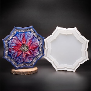 May include: A blue, flower-shaped resin coaster with a red and purple floral design and silver glitter accents. The coaster sits on a small wooden base. A white, flower-shaped silicone mold is next to the coaster.