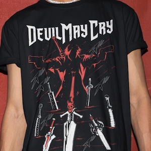 Peut inclure: T-shirt noir avec un imprimé graphique d'un personnage du jeu vidéo Devil May Cry. Le personnage tient une épée et est entouré d'autres épées. Le texte "Devil May Cry" est imprimé en blanc sur le t-shirt.