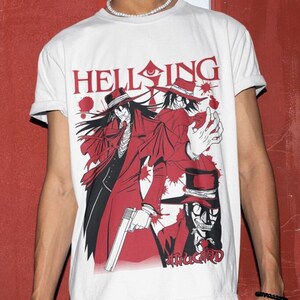 Pode incluir: Camiseta branca com uma estampa gráfica de três personagens de anime da série Hellsing. Os personagens usam roupas vermelhas e pretas e têm uma expressão ameaçadora. O texto "Hellsing" está impresso em letras vermelhas acima dos personagens.