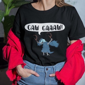 May include: Black t-shirt with a cartoon illustration of a blue creature with a beak and antlers. The creature is holding two sticks and a speech bubble above its head says "CAW CAAAW!"