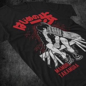 May include: Black t-shirt featuring a red and white graphic of a seated figure with wings, and Japanese text. The text "MAMORU TAKAMURA" is printed in red below the illustration. The shirt has a crew neck.