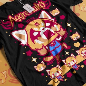 以下が含まれることがあります： 黒いTシャツ。漫画のレッサーパンダのキャラクターが描かれています。「Aggretsuko」の文字は黄色と赤色。デザインには、怒った表情のレッサーパンダと、小さなキャラクター、装飾的な要素が含まれています。
