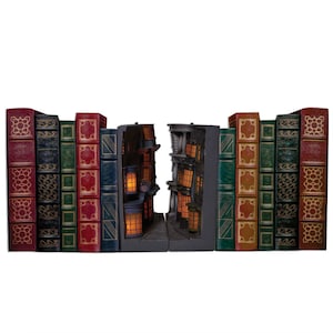 May include: A decorative bookend set featuring a miniature alleyway scene with illuminated windows nestled between rows of faux vintage books. The books have red, green, blue, and brown spines with gold embossed patterns, while the central alleyway displays dark architectural details and warm glowing lights.