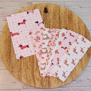 May include: A collection of dachshund-themed stationery items, including bookmarks and notepads, featuring pink and red hearts and dachshunds in sweaters. The items are arranged on a wooden cutting board with a tree design.