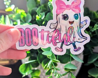 May include: A holographic sticker featuring a cartoon ghost with a pink bow, shoes, and the word "BOOtiful" in pink and purple. The ghost has large eyes and is smiling. The sticker is held in front of a background of green plants.