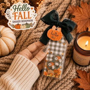 May include: A clear shaker bookmark featuring a black and white buffalo check pattern, filled with small pumpkin and flower-shaped confetti. It is topped with a dark green fabric bow and an orange pumpkin charm with the text hello FALL.