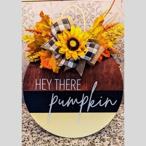 May include: A round wooden door hanger with a black and white checkered ribbon, a yellow sunflower, and fall leaves. The sign reads "Hey there pumpkin".