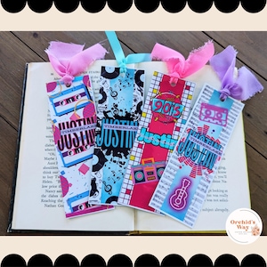 May include: Four colorful bookmarks with a 90s theme. Each bookmark features a different design with a cassette tape, a boombox, a guitar, and a music note. The bookmarks are decorated with a pink, blue, and purple ribbon.