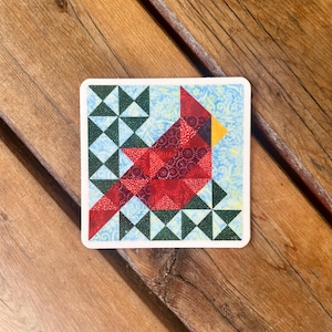 May include: A red cardinal bird made from fabric squares, with a yellow beak, is the focal point of this colorful patchwork design. The background is a light blue with green and white geometric shapes.