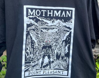 Puede incluir: Camiseta negra con un gráfico blanco. El gráfico incluye la palabra "MOTHMAN" sobre una ilustración detallada de un puente, montañas, árboles y una criatura. El texto "POINT PLEASANT" está debajo.