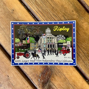 May include: A sticker featuring a colorful illustration of a town square with a courthouse, fireworks, and a parade. The text "Ripley, Est. 1832, USA's Largest Small Town Independence Day Celebration" is printed on the sticker.