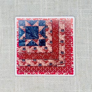 May include: Square sticker featuring a quilt design with a star pattern. The quilt has a red, white, and blue color scheme with floral and leaf patterns. The sticker is square with a white border and is placed on a textured, neutral background.