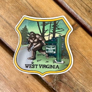 May include: A sticker featuring a green and yellow shield with a cartoon Bigfoot walking on a path in a forest. The text "Greenbrier State Forest" is on a signpost in the image. The text "West Virginia" is at the bottom of the shield.
