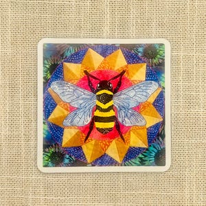 May include: A square sticker featuring a detailed illustration of a bee. The bee has a black and yellow striped body, blue wings, and is centered on a colorful geometric design with gold, red, and blue hues. The sticker is bordered in white.